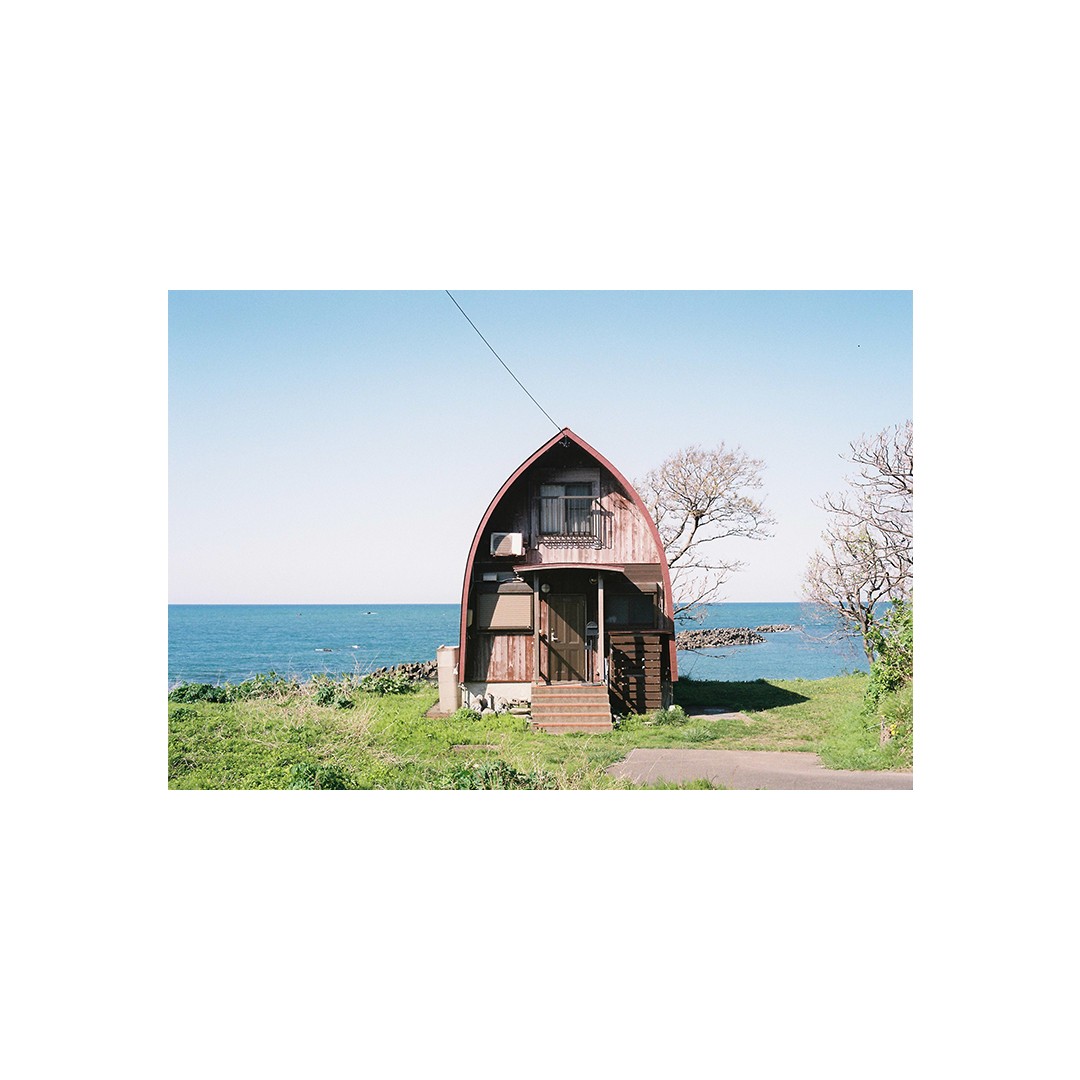 A small, boat-shaped wooden house stands on a grassy hill overlooking the ocean under a clear sky.