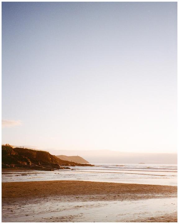 A sandy beach with waves crashing on the shore next to a rocky cliff under a clear sky.