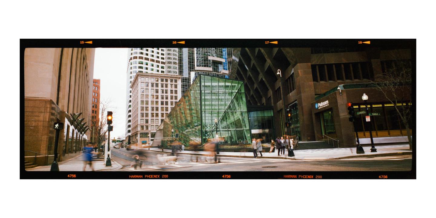 A street scene shows a modern glass building and other city buildings with blurred people and vehicles moving along the street.