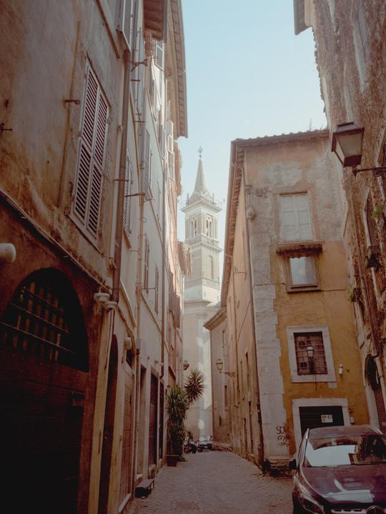 A narrow cobblestone street with tall buildings on either side leads to a distant bell tower under a clear sky.