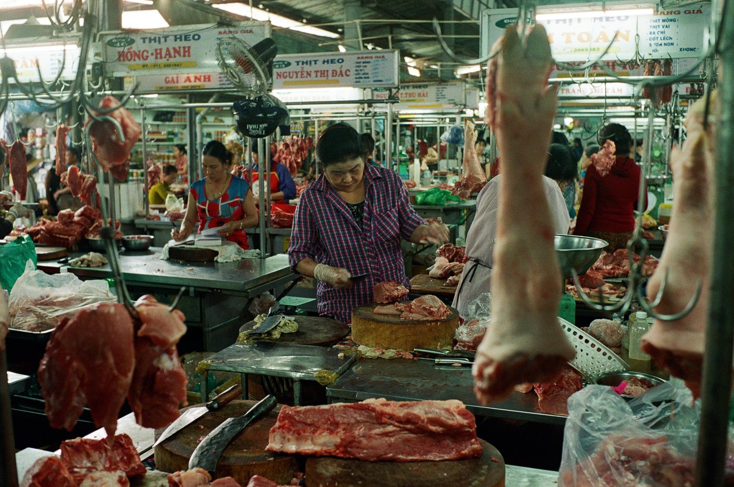 A woman in a plaid shirt cuts meat at a busy market stall with various cuts of meat displayed.