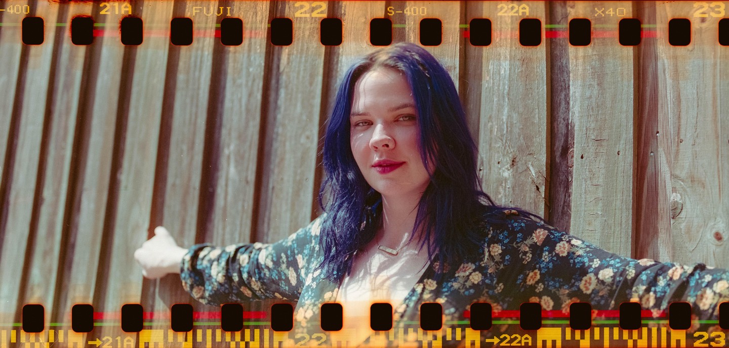 A woman with blue hair and a floral shirt stands in front of a wooden fence.