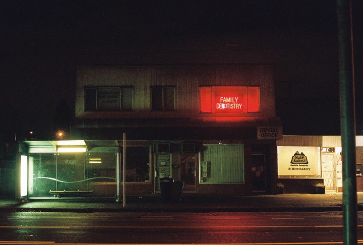 A street scene at night shows a bus stop, a dental office with a red neon sign, and a bakery.