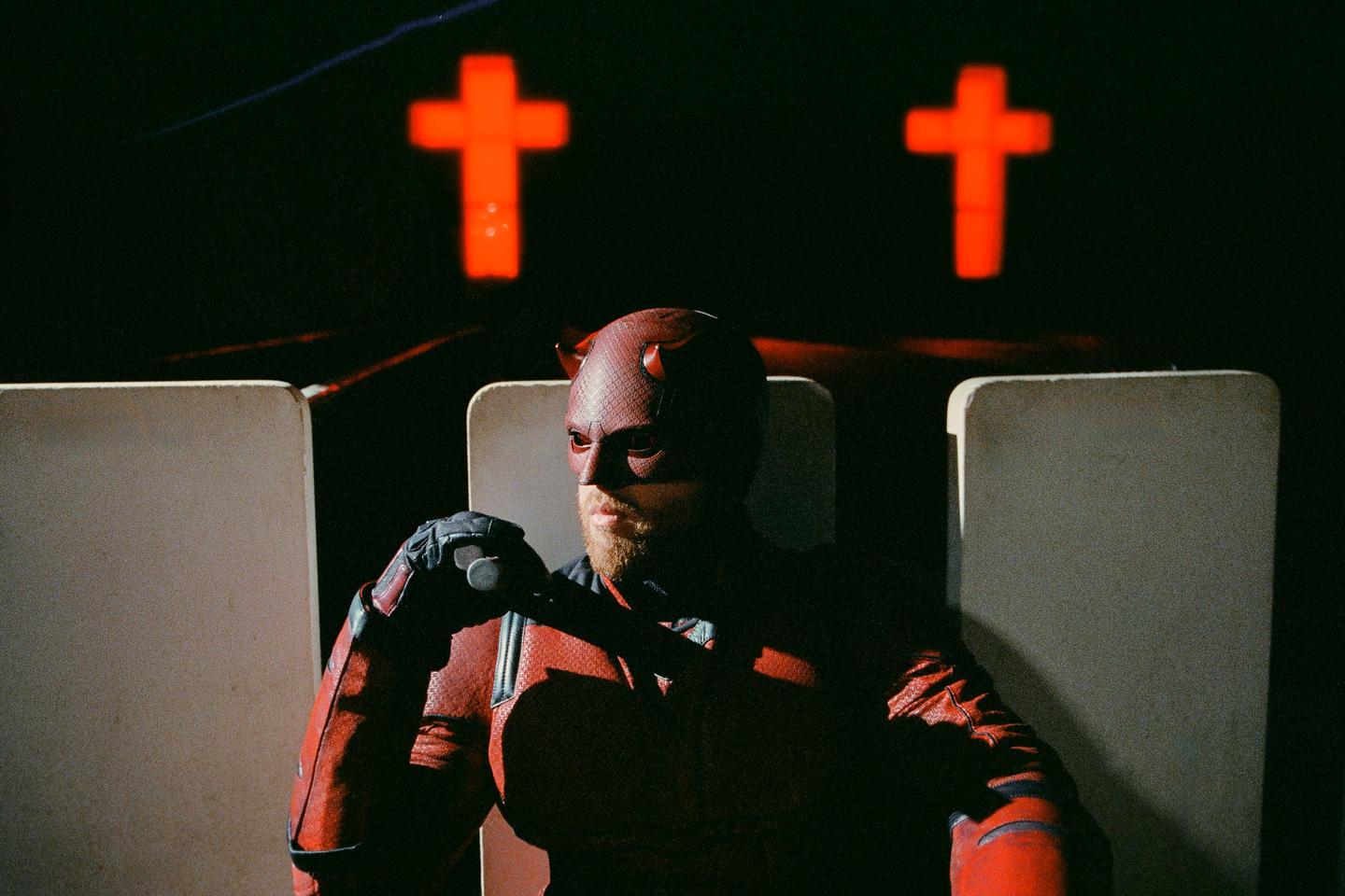 A man in a red Daredevil costume sits in front of two glowing red crosses.