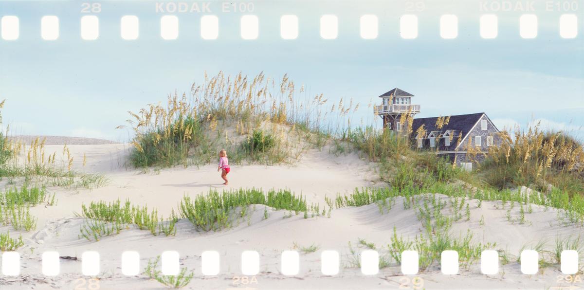 A child walks on a sandy dune with a house and watchtower in the background.