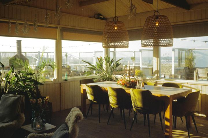 A dining room with a long wooden table and green chairs is illuminated by sunlight streaming through large windows overlooking a beach.