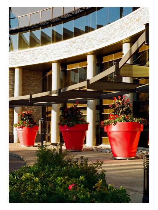 Three large red planters with flowers sit in front of a modern building with reflective windows.