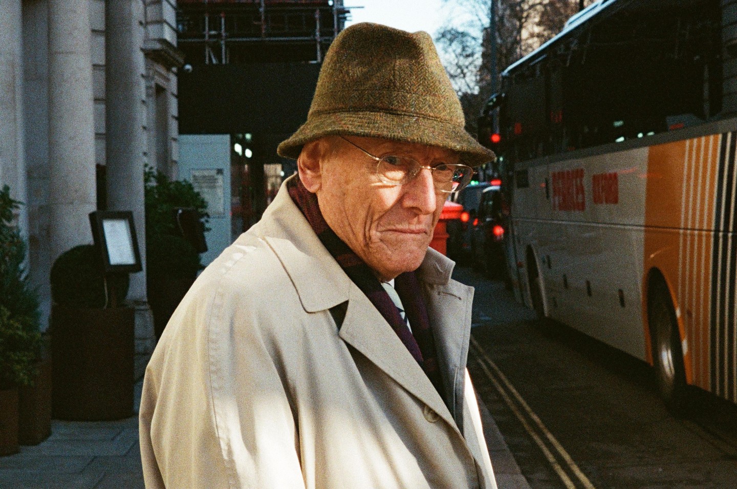 A man in a trench coat and hat stands on a street next to a bus.