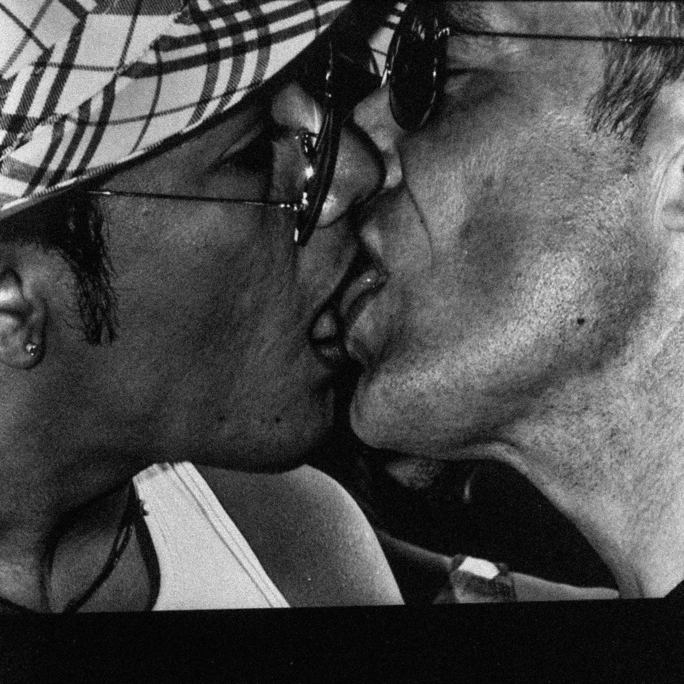 Two men kiss each other, one wearing a plaid hat and sunglasses, the other wearing sunglasses.