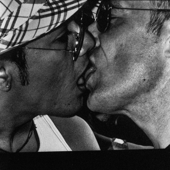 Two men kiss each other, one wearing a plaid hat and sunglasses, the other wearing sunglasses.
