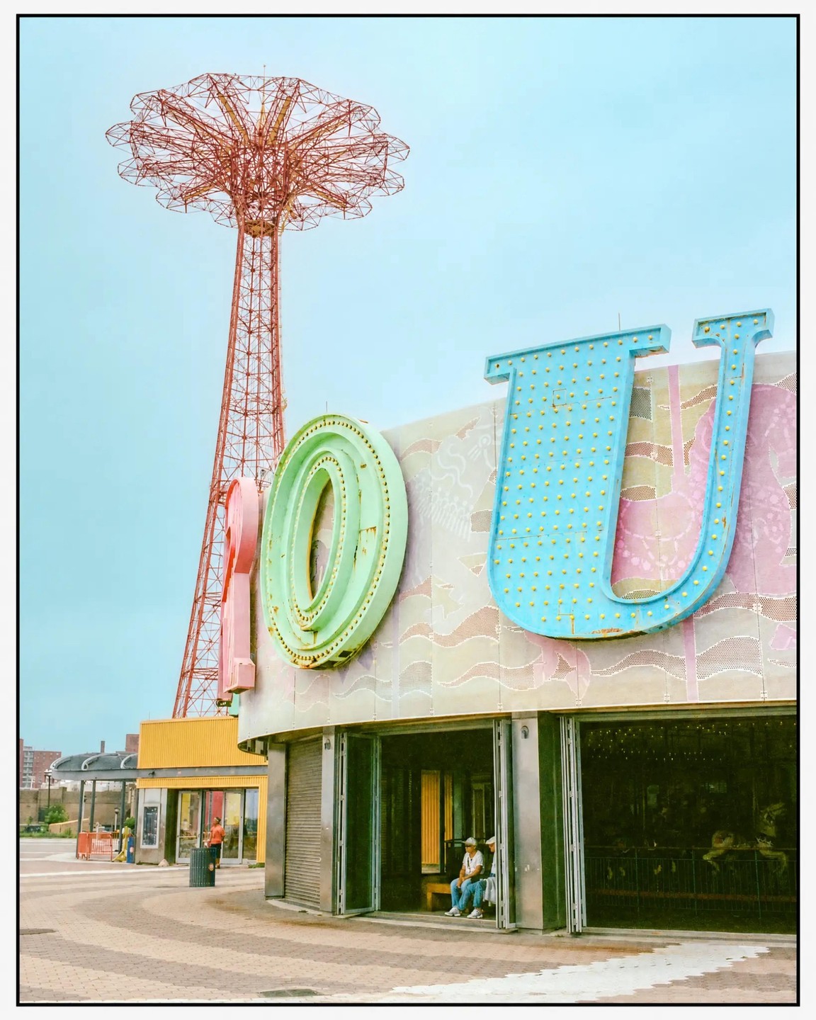 A large red parachute jump ride stands tall behind a building with colorful letters spelling "YOU" on its facade.