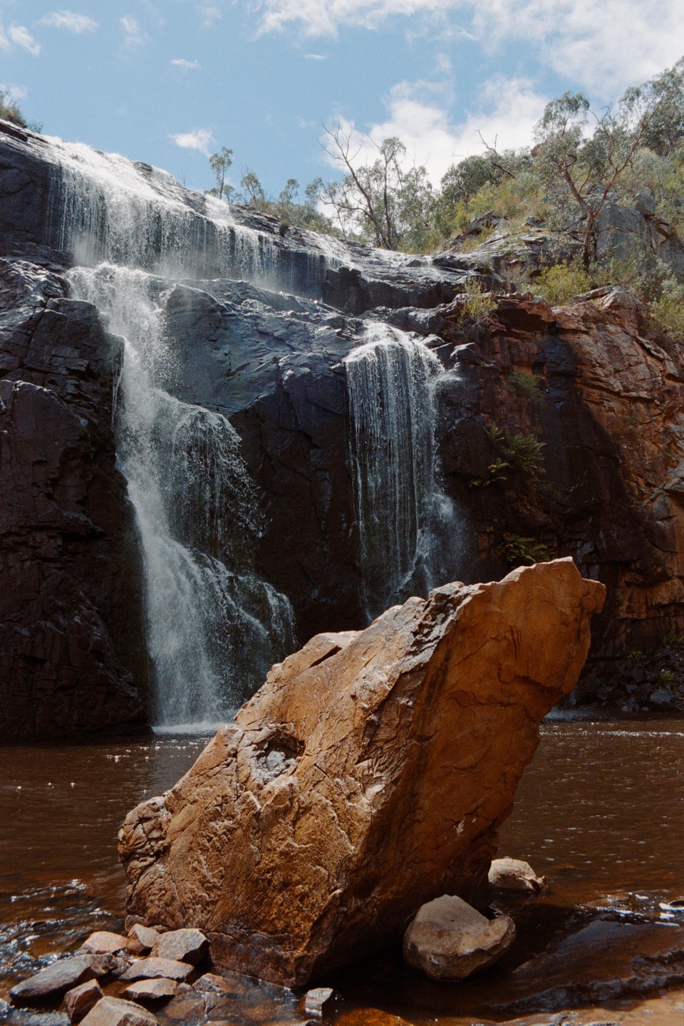A large waterfall cascades over dark rocks into a brown pool, with a prominent orange boulder in the foreground.