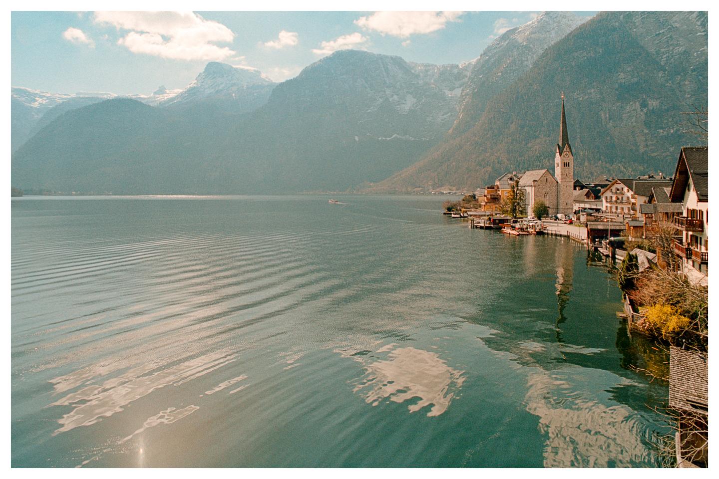 A village with a tall church steeple sits on the edge of a lake, surrounded by mountains.