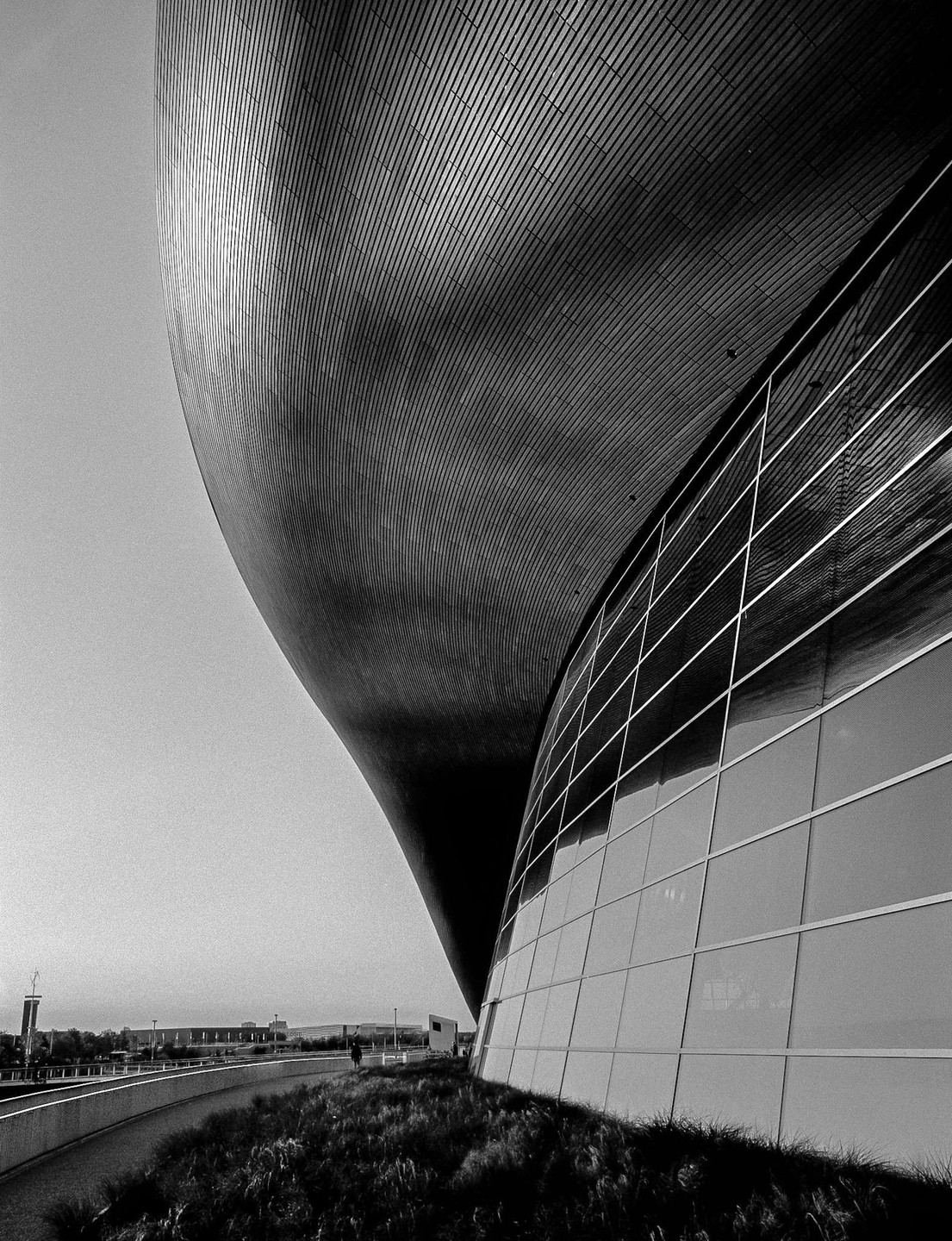 A large, curved building with a textured roof and glass facade stands next to a grassy area and a walkway.