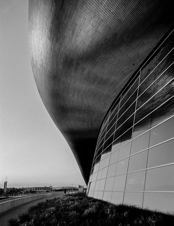 A large, curved building with a textured roof and glass facade stands next to a grassy area and a walkway.