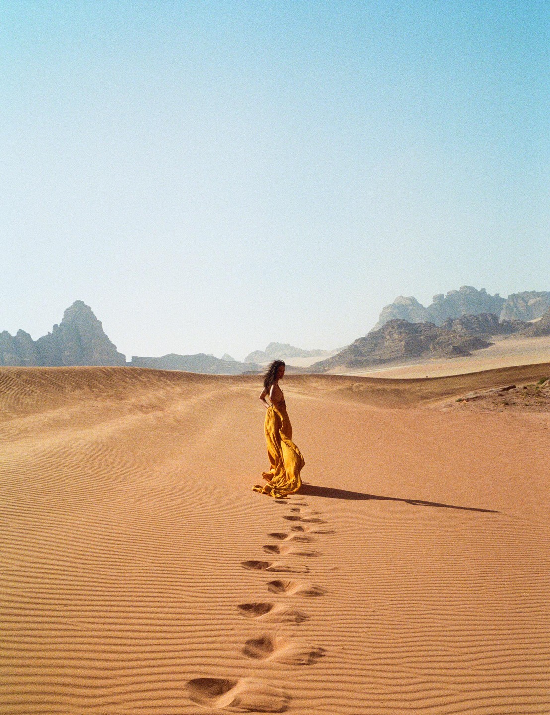 A woman in a flowing yellow dress stands in a vast desert with footprints leading away from her.