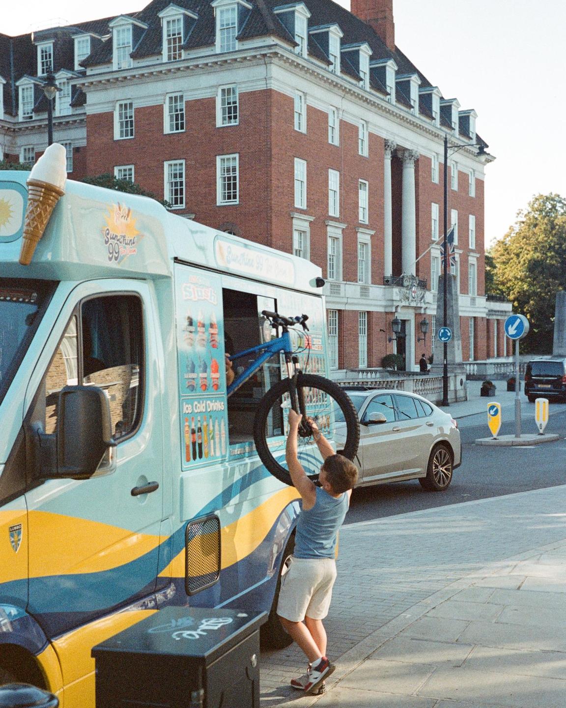 A boy holds a bicycle wheel while standing next to an ice cream truck on a street in front of a large brick building.
