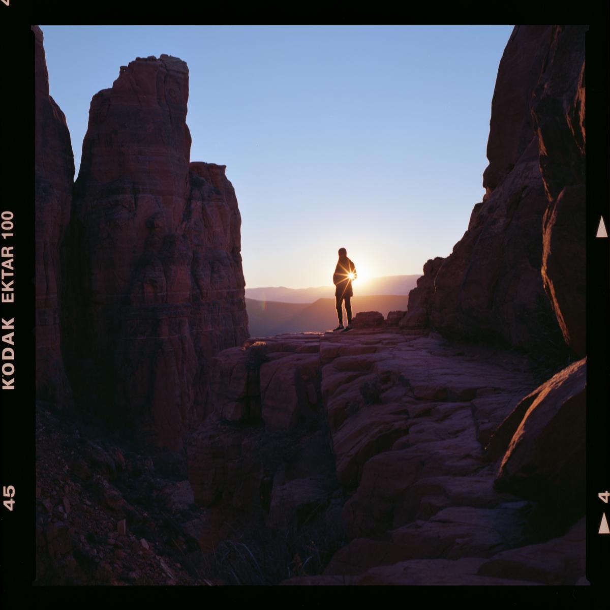 A person stands on a rock formation at sunset, with mountains in the distance.