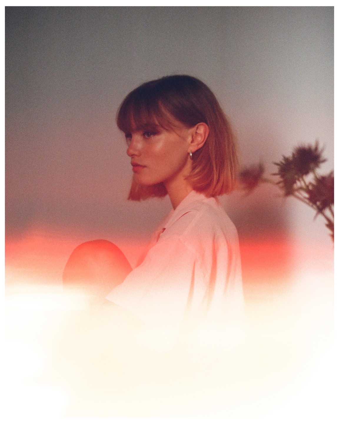 A woman with short hair and bangs looks to the left, with a plant visible on the right.