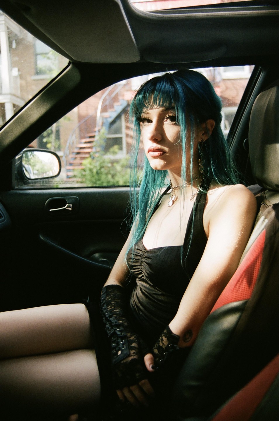 A woman with blue hair and a black dress sits in a car.