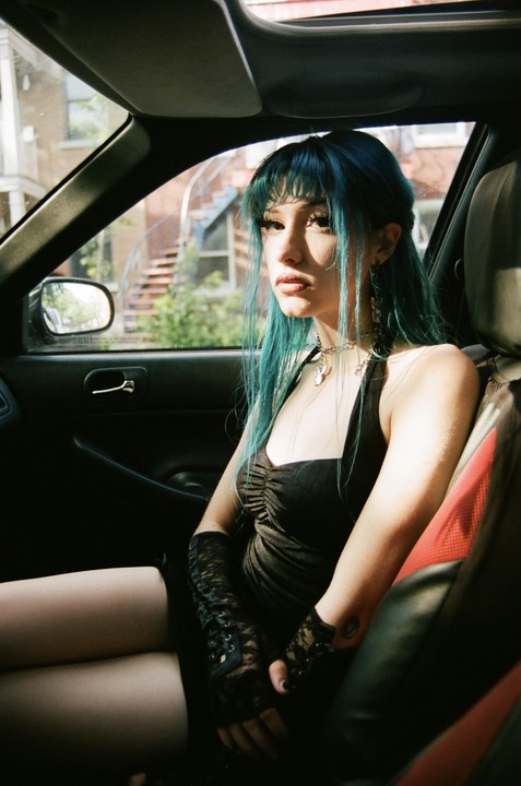 A woman with blue hair and a black dress sits in a car.