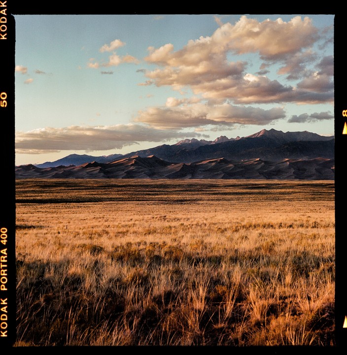 A vast field of dry grass stretches towards sand dunes and mountains under a cloudy sky.