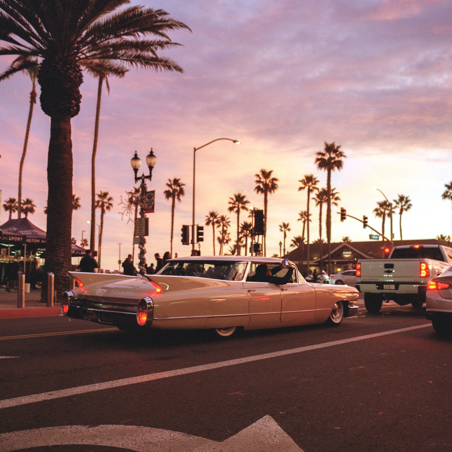 A vintage car drives down a street lined with palm trees at sunset.