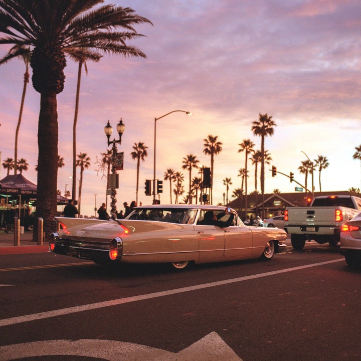 A vintage car drives down a street lined with palm trees at sunset.