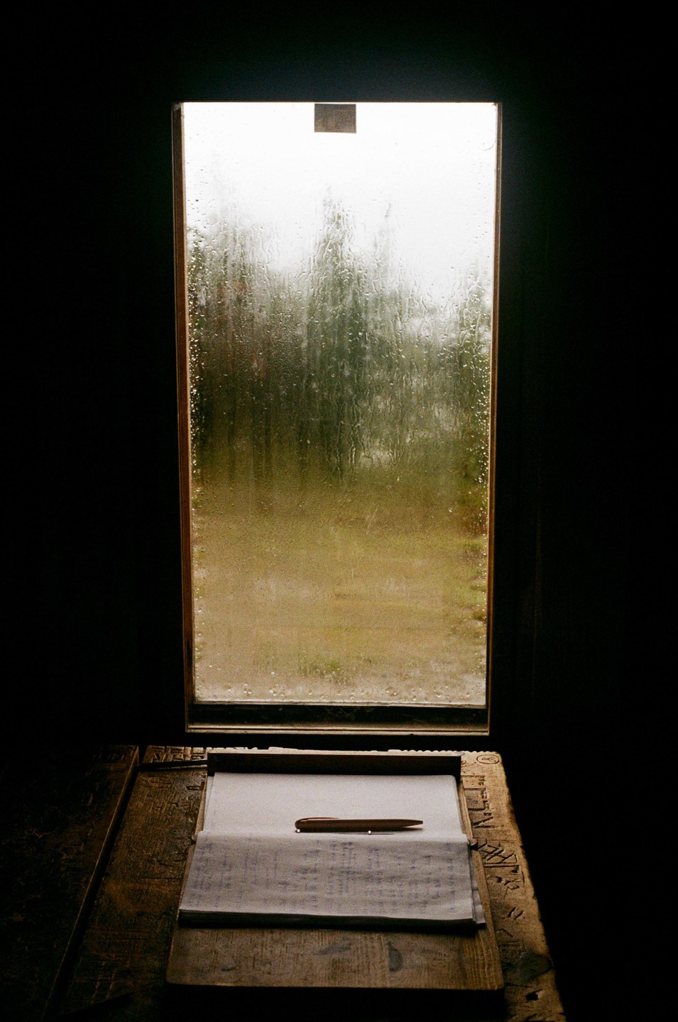 A pen rests on a piece of paper on a wooden desk in front of a window covered in rain.