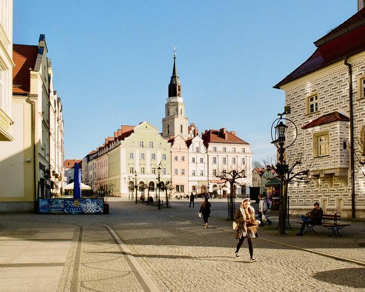 A woman walks across a cobblestone town square with historic buildings and a tall clock tower under a clear sky.