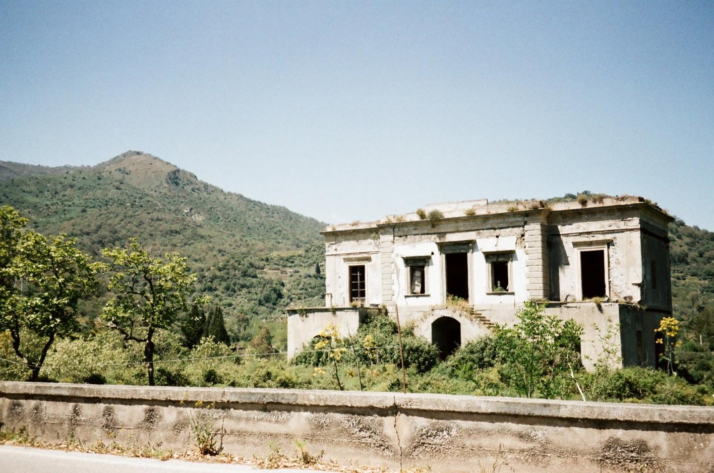An abandoned building with overgrown vegetation stands before a mountain under a clear sky.