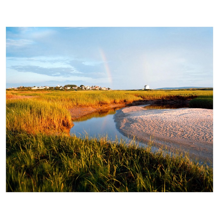 A rainbow stretches over a coastal landscape with a winding tidal creek, marsh grass, and a distant town with a lighthouse.