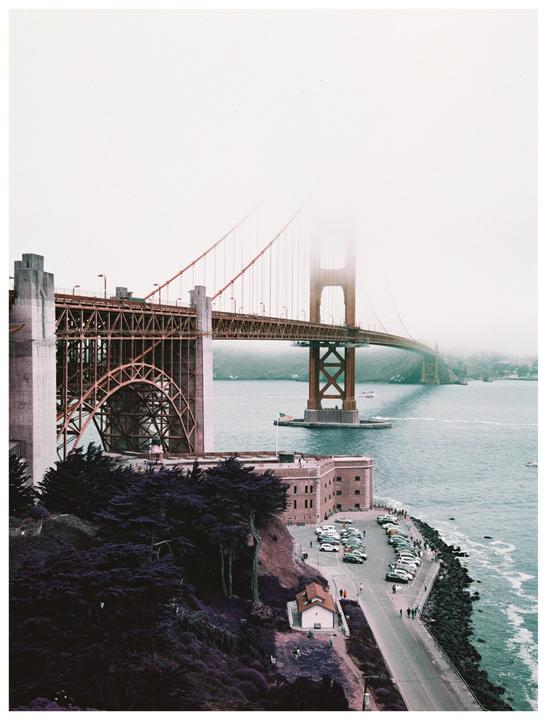 The Golden Gate Bridge is partially obscured by fog, with a building and parking lot visible on the shore below.