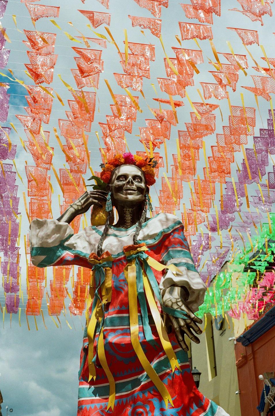 A large skeleton figure wearing a colorful dress and flower crown stands beneath a canopy of papel picado.