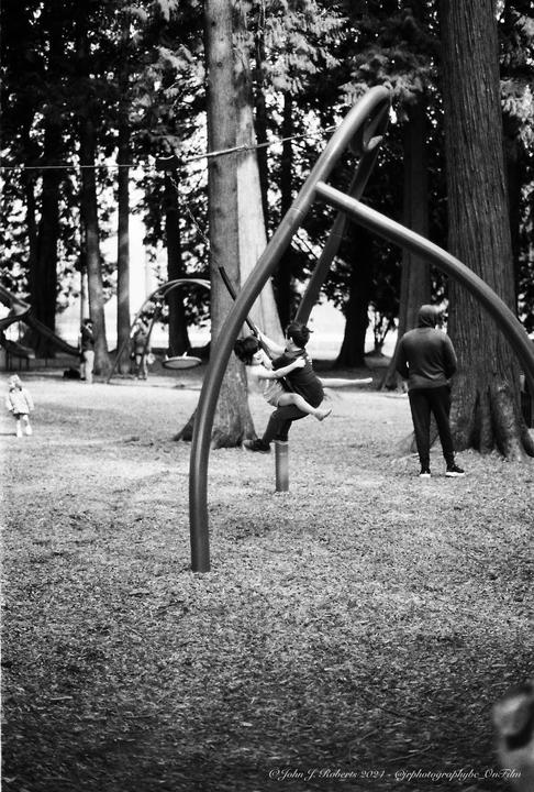 Two children play on a swing set in a park while an adult stands nearby.