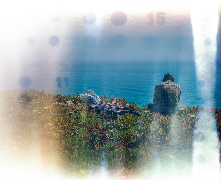A person sits on a grassy cliff overlooking the ocean, with a blanket and a red bottle beside them.