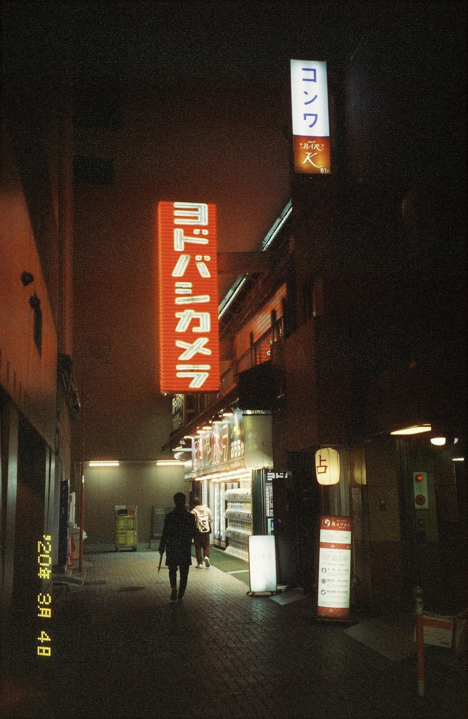 A person walks down a narrow street at night, illuminated by neon signs and storefront lights.