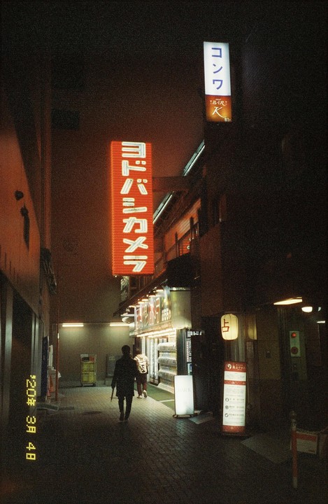 A person walks down a narrow street at night, illuminated by neon signs and storefront lights.