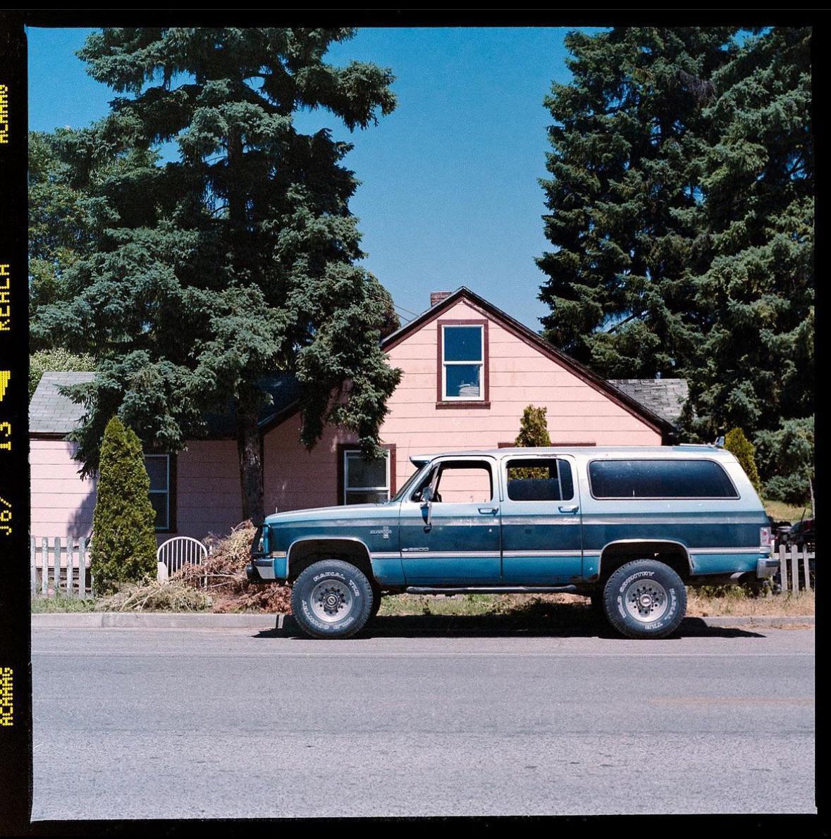 A blue and white SUV is parked on the side of a road in front of a pink house with tall evergreen trees.