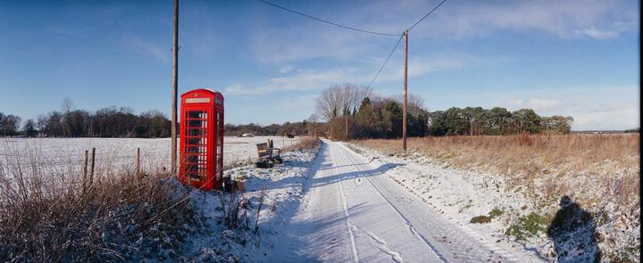A red telephone booth stands next to a snow-covered road with fields and trees under a clear sky.