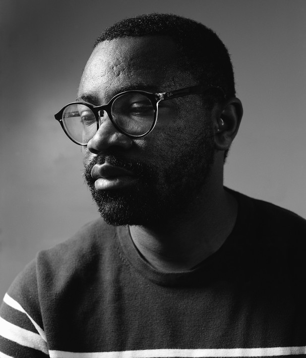 A man wearing glasses and a striped sweater looks down and to the side in a black and white portrait.