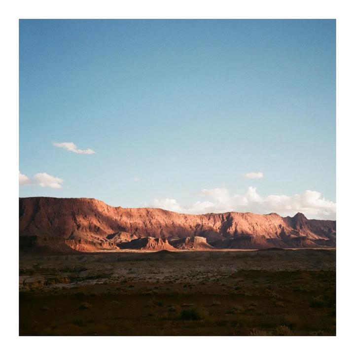 A vast desert landscape features a long, red rock formation under a clear blue sky with scattered clouds.