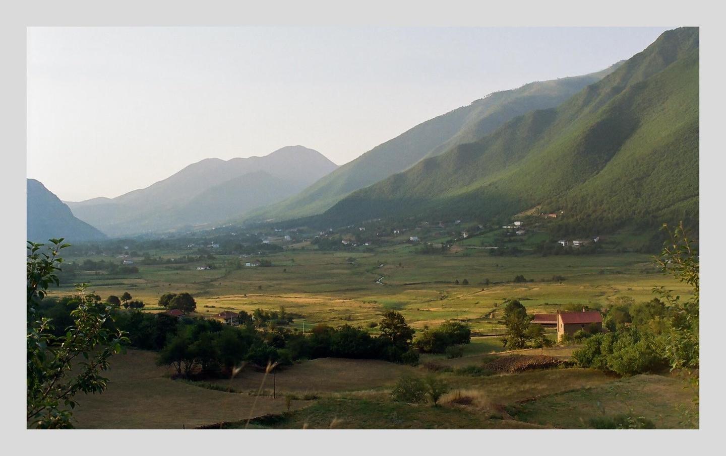 A valley with a small village and a single house is nestled between large mountains under a hazy sky.