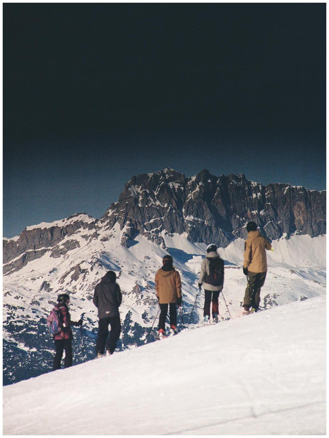 Four skiers stand on a snowy slope looking at a large mountain range under a dark sky.