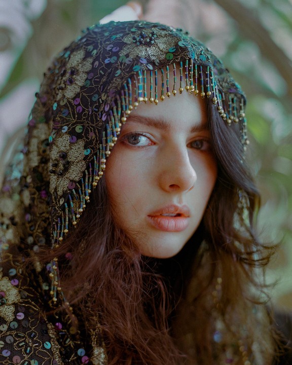 A woman with long, dark hair wears an ornate, beaded hood with sequins.