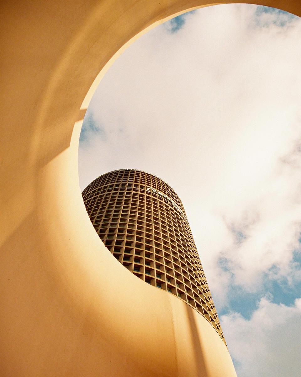A tall, cylindrical building with a grid pattern on its facade is framed by a curved, light-colored structure against a cloudy sky.