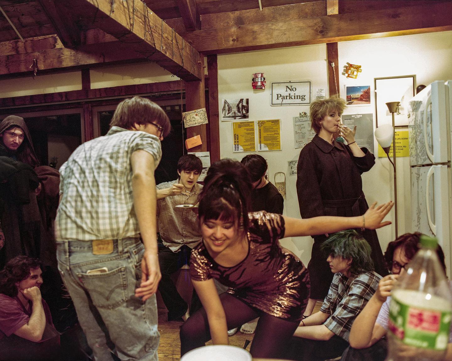A group of young adults are gathered in a dorm room, some standing and some sitting on the floor, with one woman smoking a cigarette.