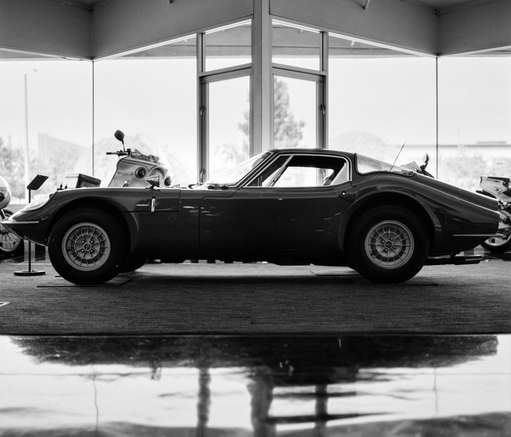 A vintage sports car is parked on a carpeted floor with a large window behind it.