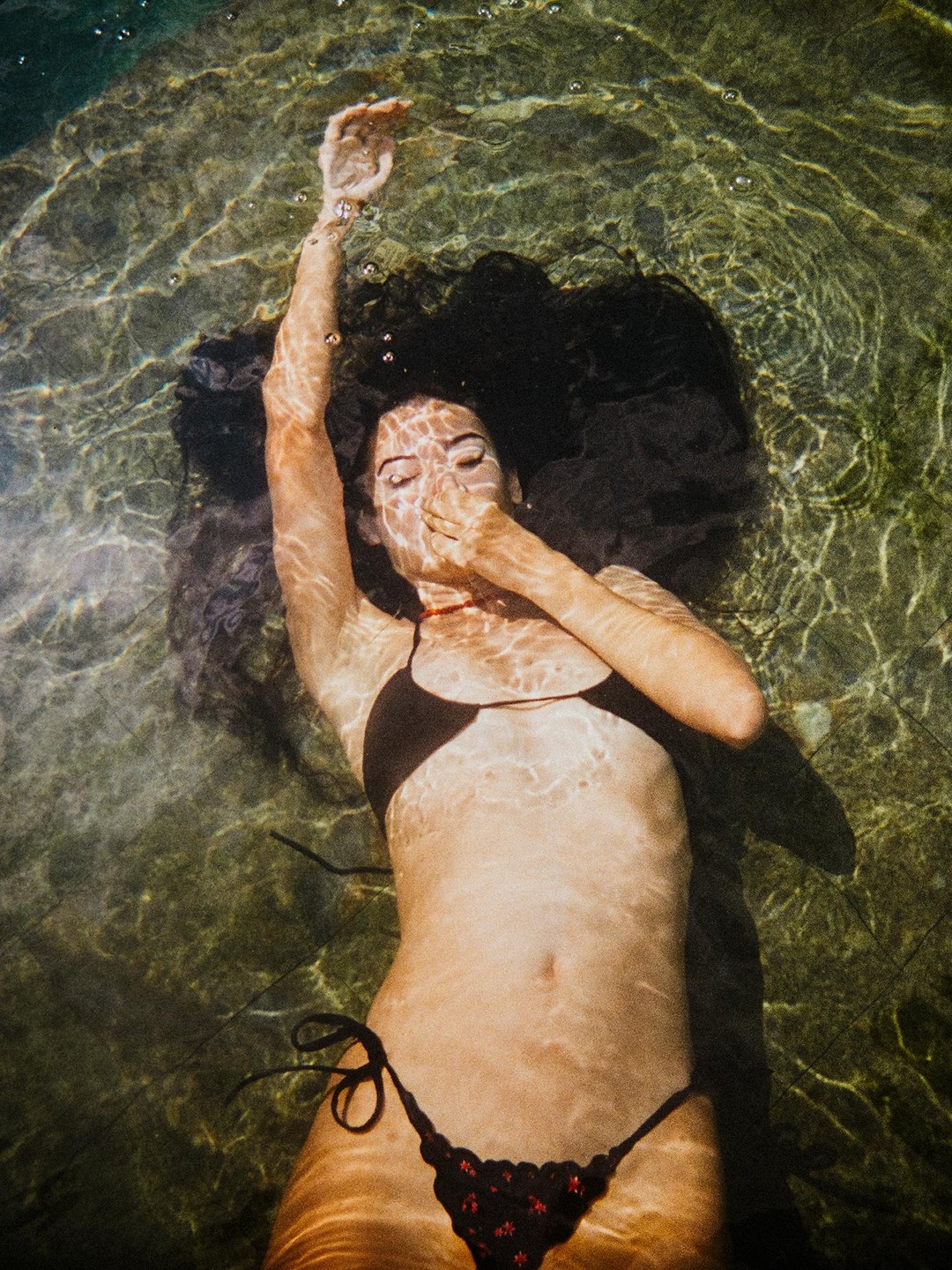 A woman in a bikini floats on her back in clear water, with her hand covering her mouth and her other arm raised.