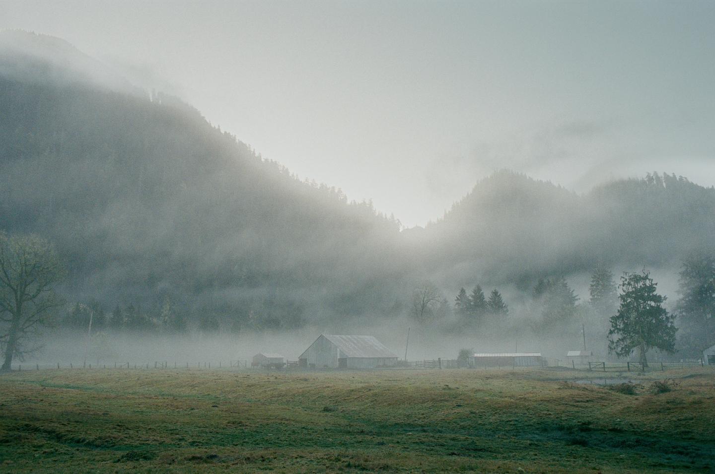 A misty morning view shows a barn and field in a valley surrounded by forested mountains.